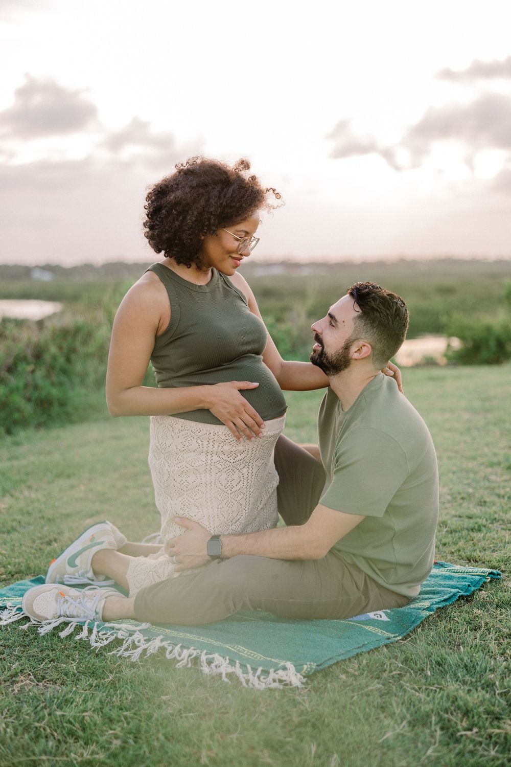A couple sits together on a turquoise blanket in a grassy field during sunset sharing an intimate moment.