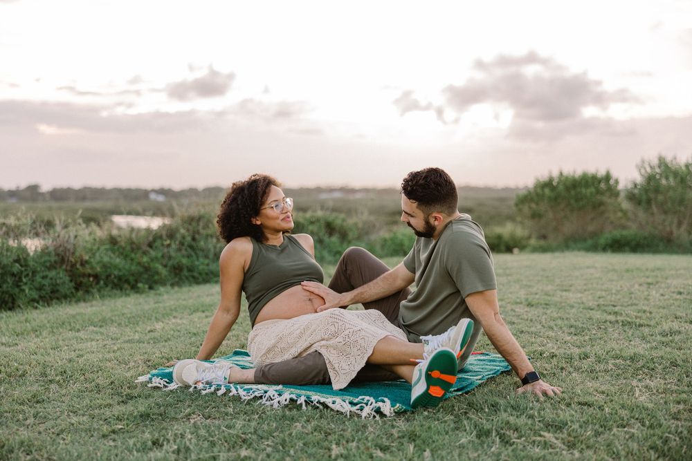 A couple sits together on a blanket in a grassy field during sunset, sharing an intimate moment outdoors.