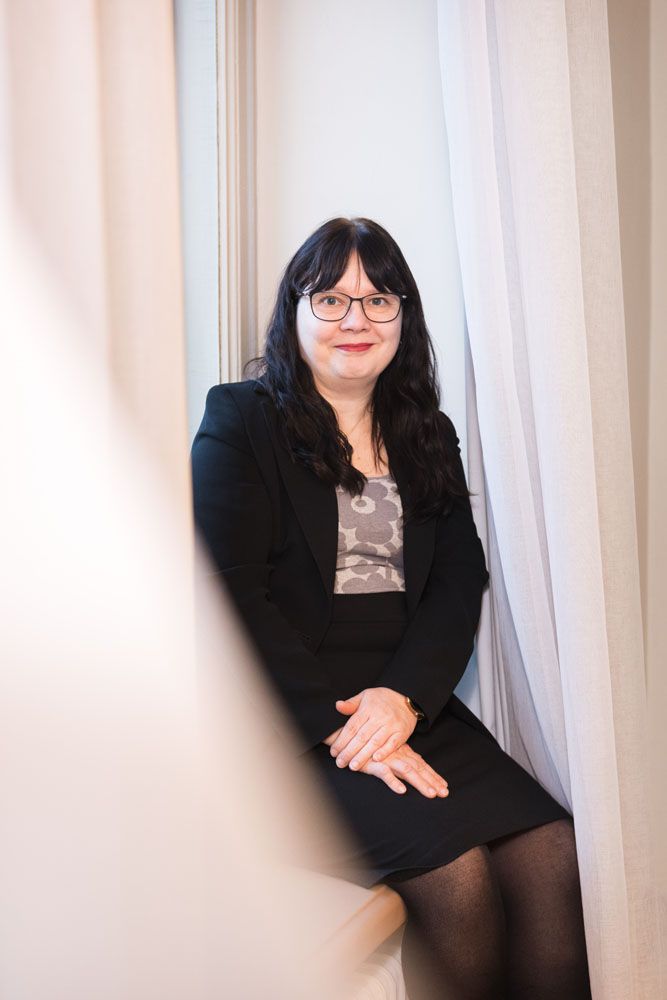 50-vuotias mustahiuksinen nainen istuu ikkunalaudalla ja katsoo kameraan lempeästi hymyillen.