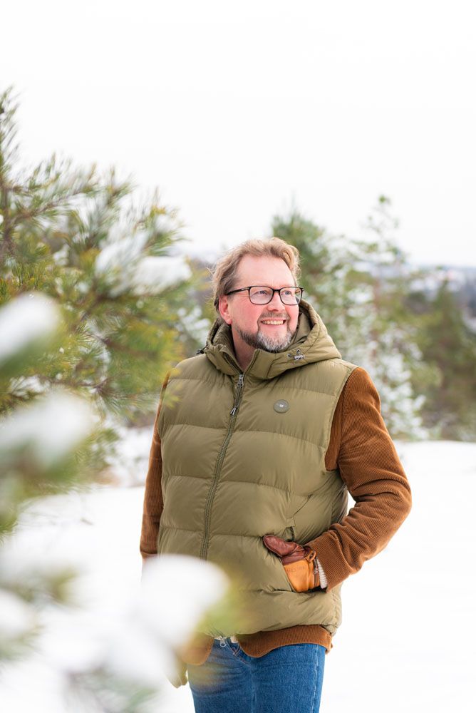 60-vuotias mies seisoo talvimaisemassa hymyillen ja päällään vihreä liivi ja siniset farkut.