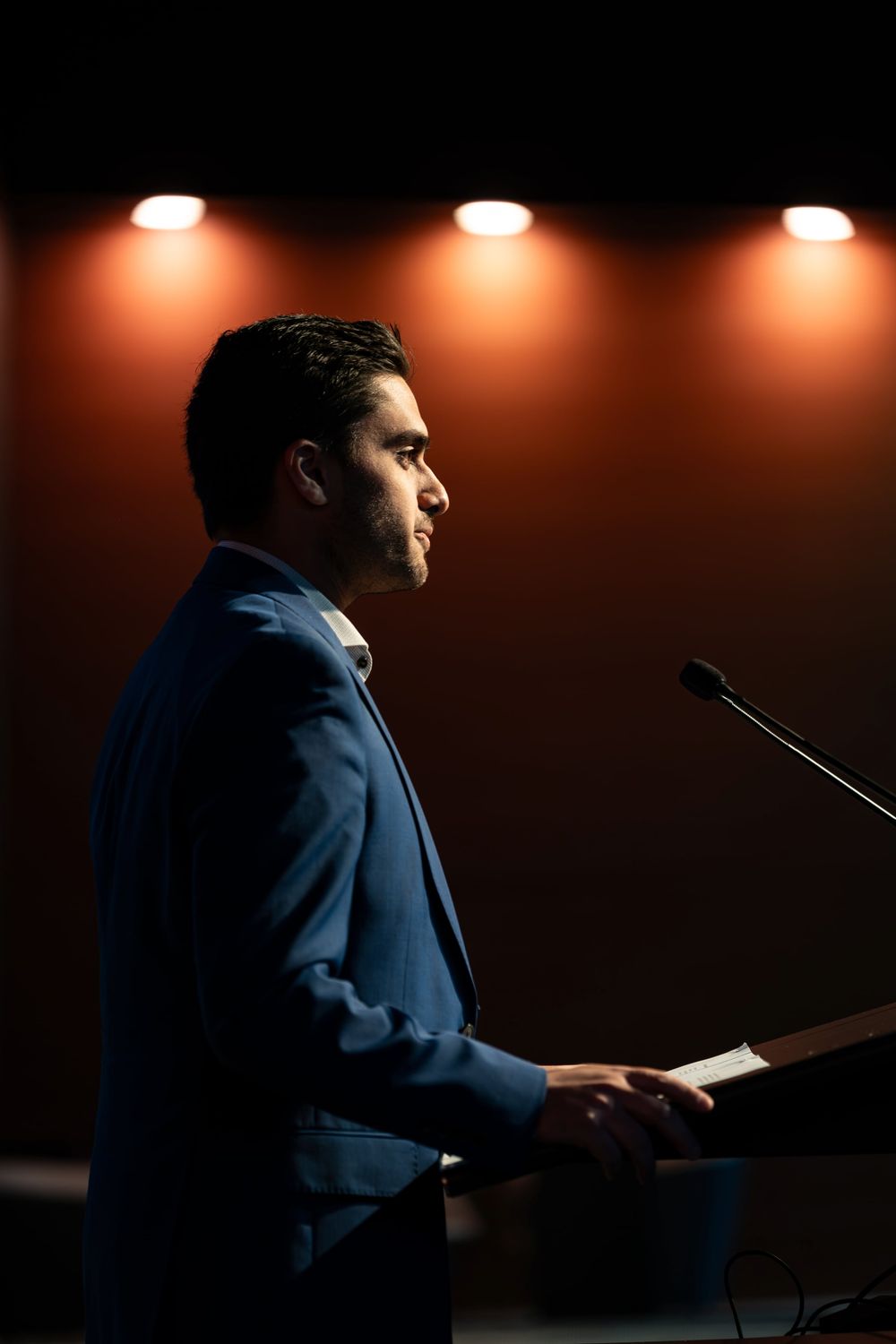 Businessman in blue suit speaking at podium under warm stage lights.