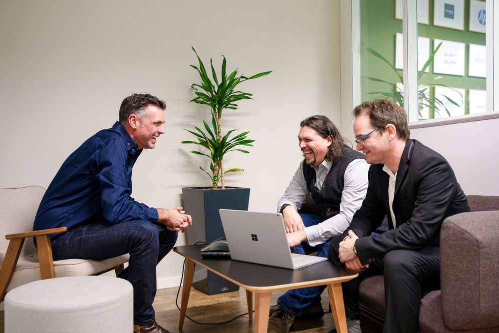Three professionals laughing and collaborating on a laptop in a modern office lounge area.