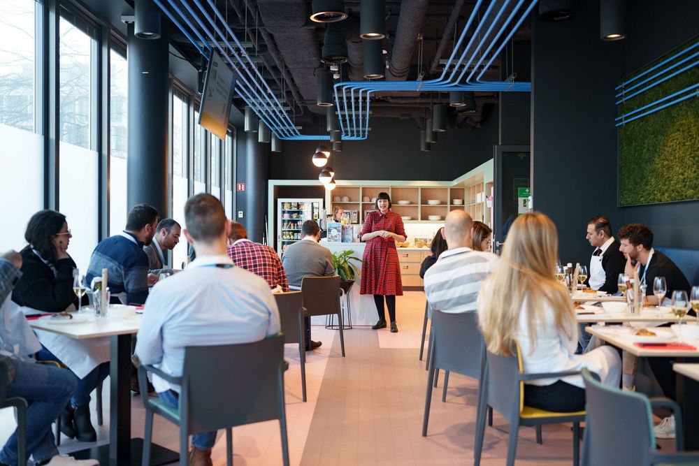 People dining in a modern restaurant while a presenter in red dress addresses the seated guests.
