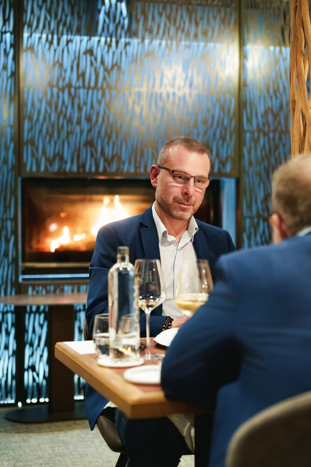 Man in blue blazer having a business dinner conversation by a fireplace in upscale restaurant.