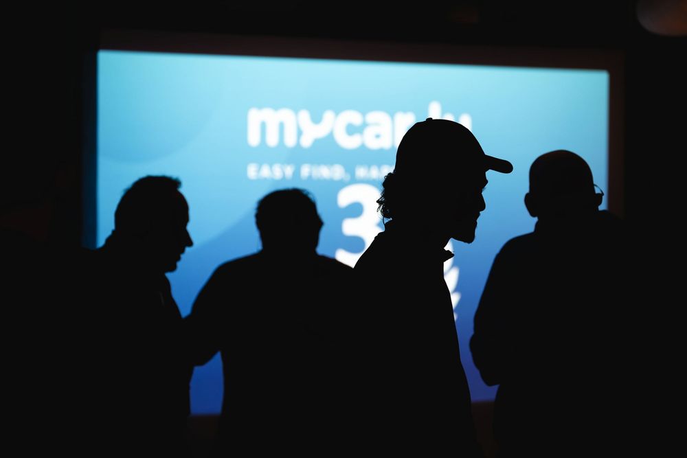 Silhouettes of people at a MyCar3 product launch event with blue projection screen backdrop.