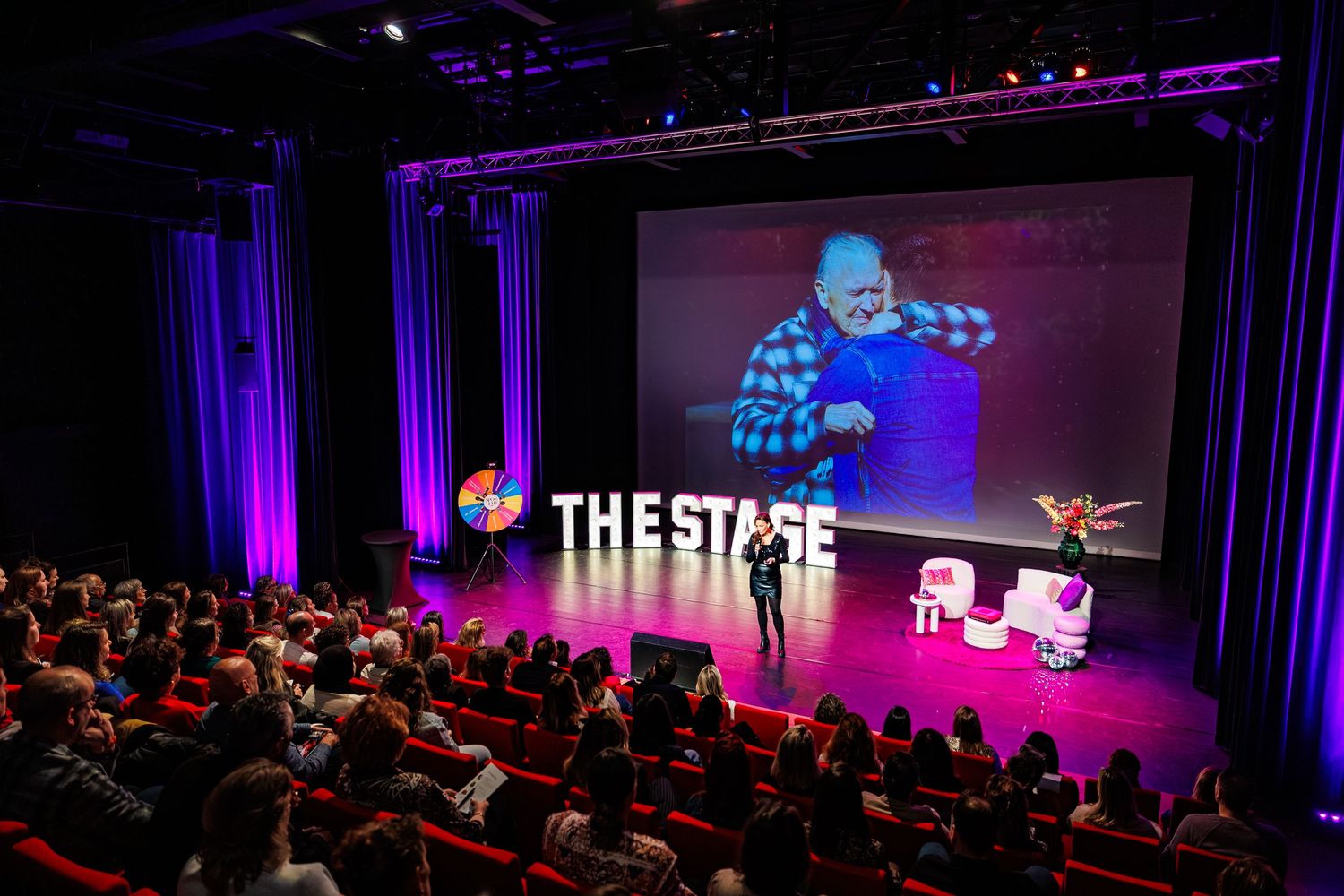 Audience members seated in a dark theater with purple stage lighting illuminating the presentation space.