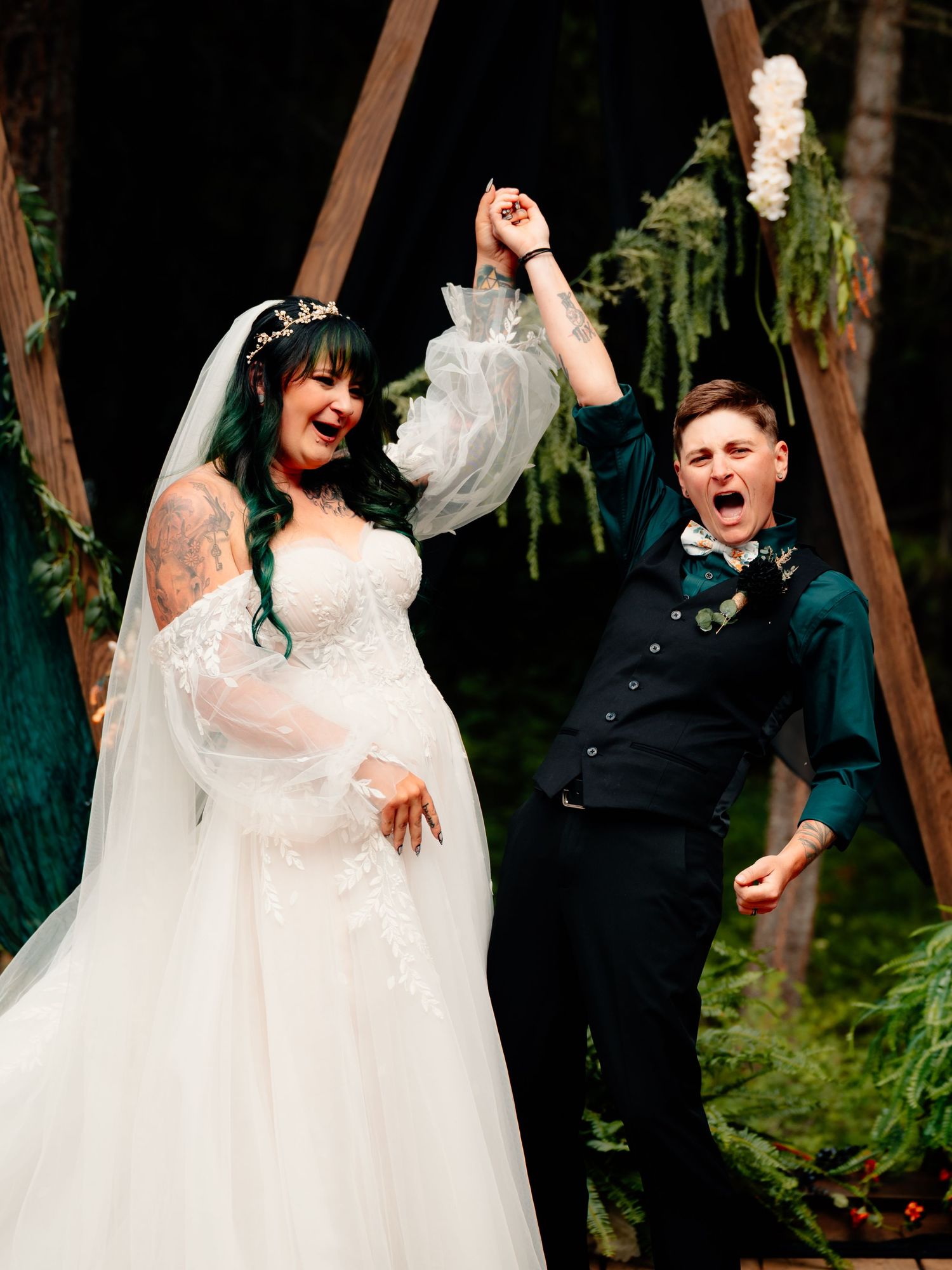 Wedding couple celebrates joyfully with raised arms during reception at outdoor venue with wooden beams and greenery.