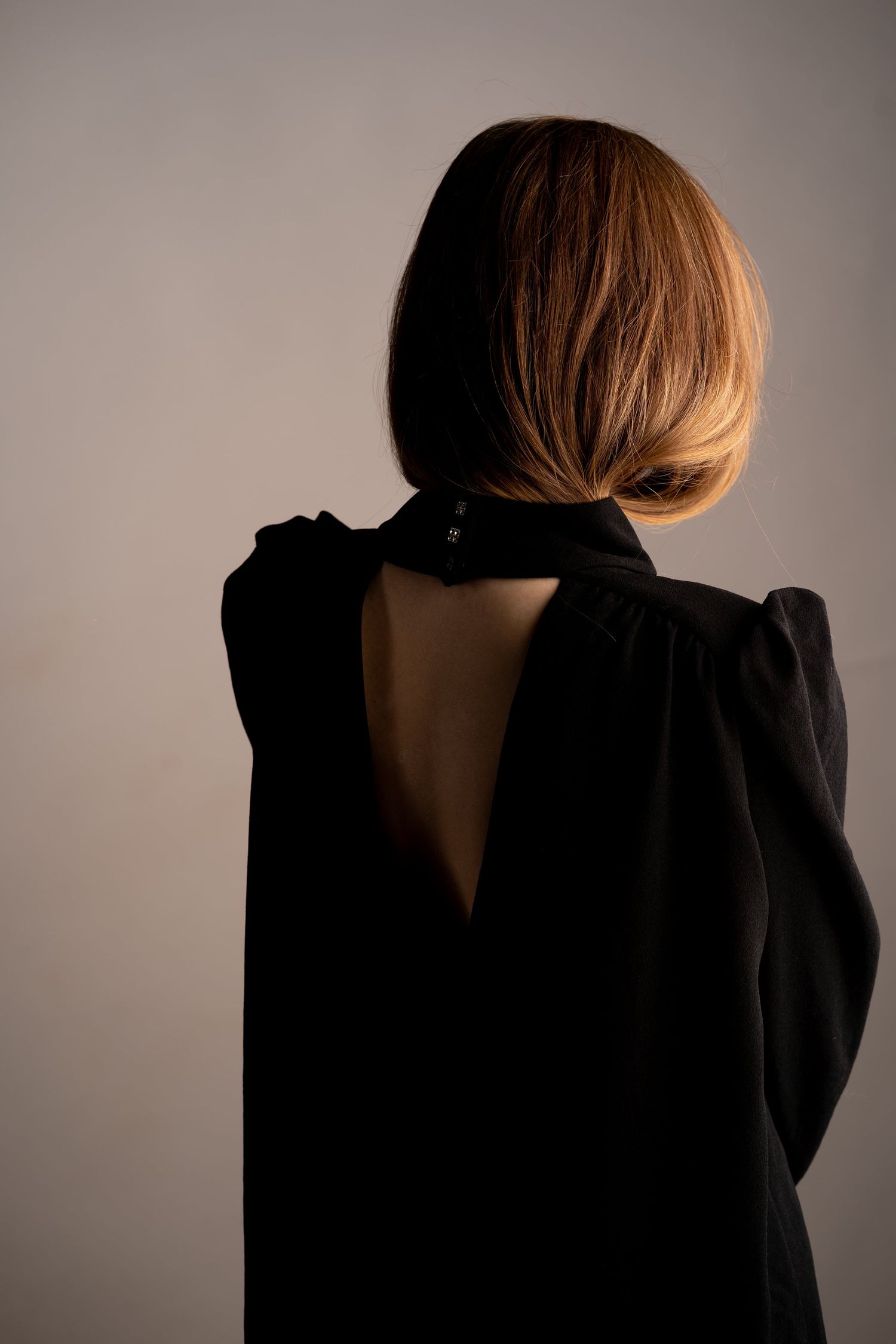 Woman in black open-back dress with bob haircut photographed from behind on neutral background.