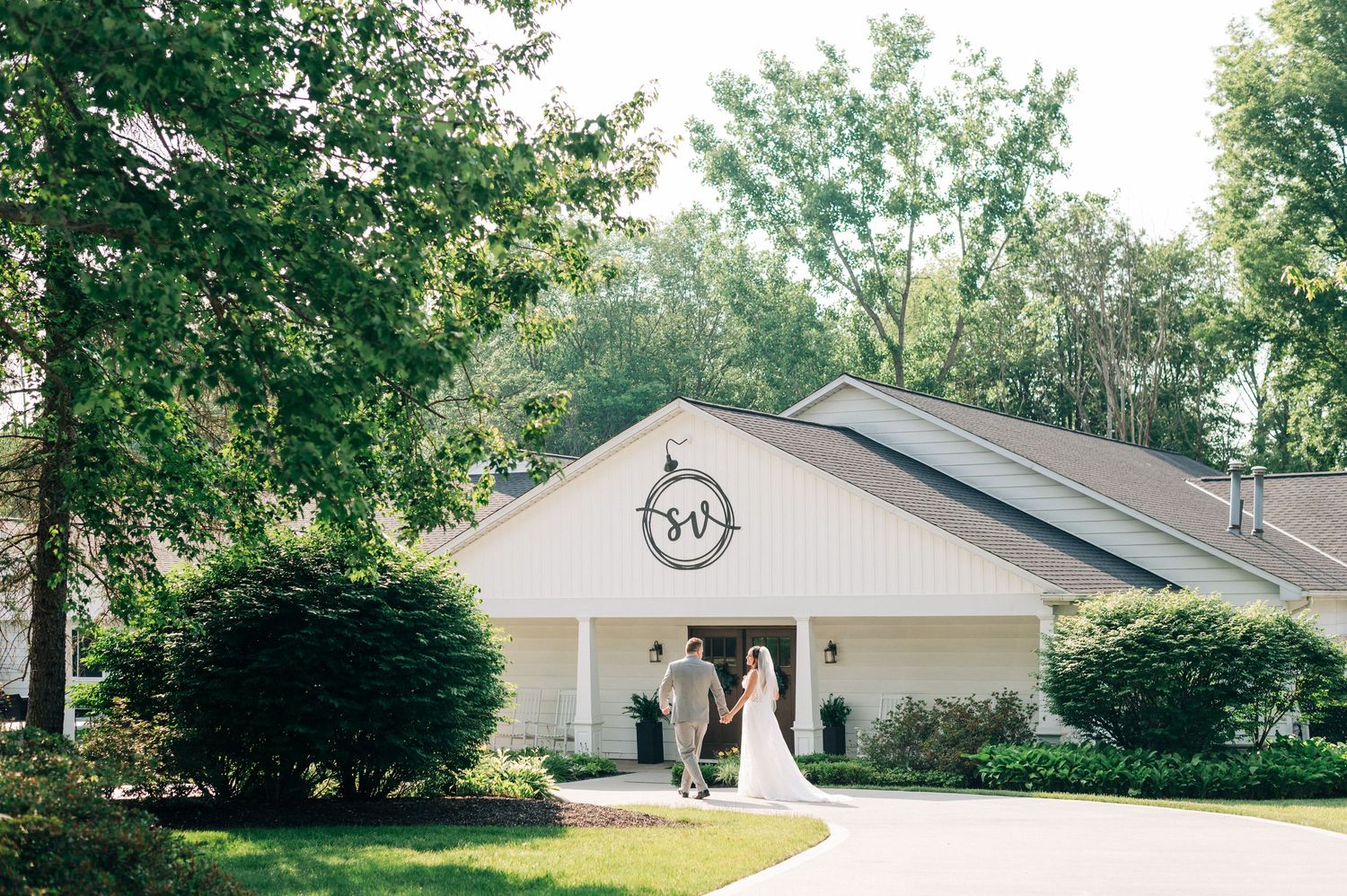 A white ranch-style house with attached garage and landscaping sits nestled among green trees on a sunny day.