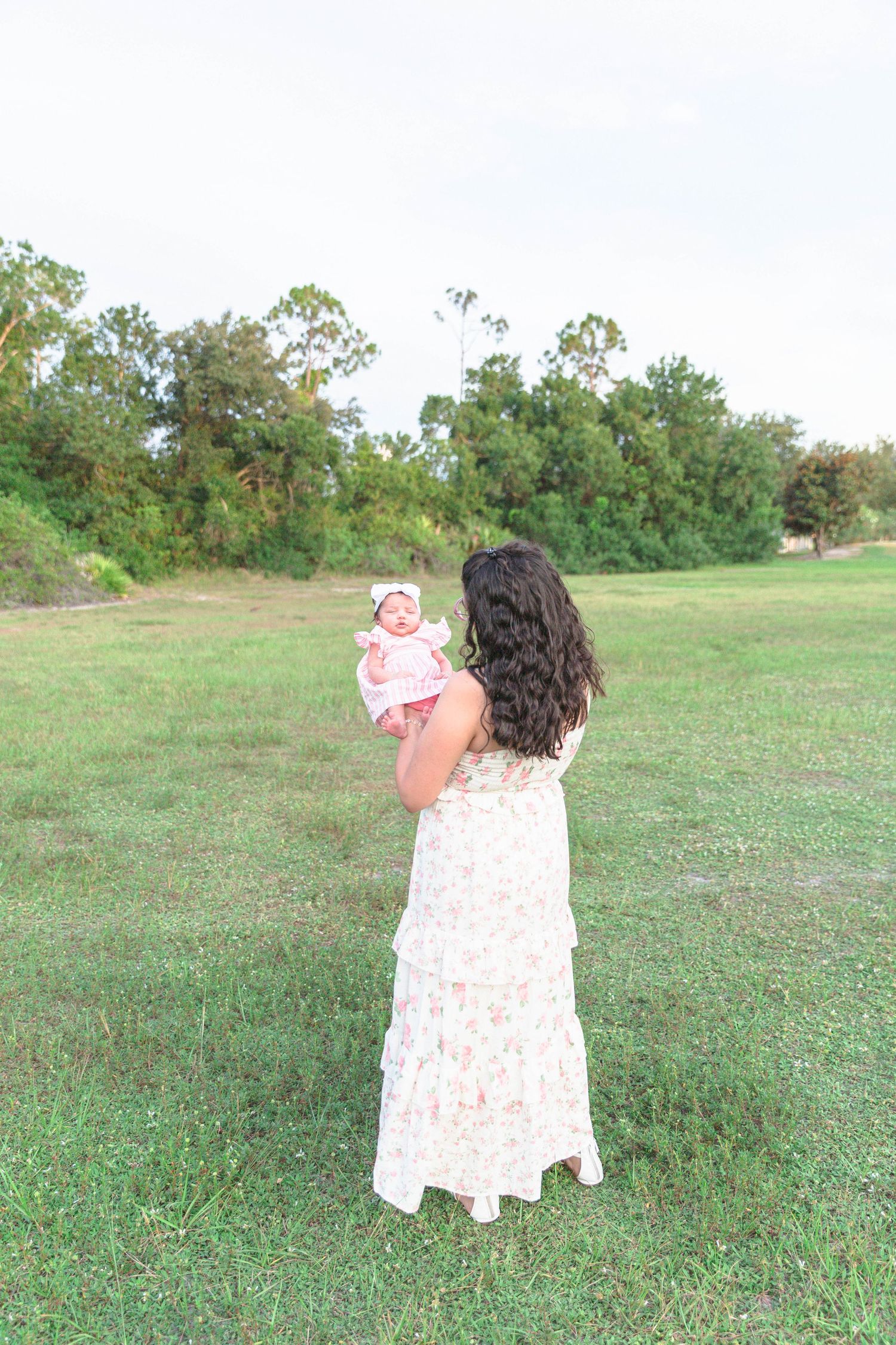 Dark-haired mother in floral dress holding her infant baby girl up in a green meadow.