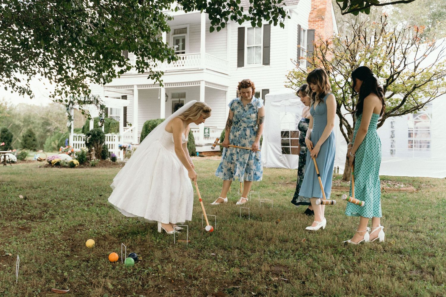 Wedding party in blue dresses and white gowns playing croquet on a lawn during an outdoor celebration.