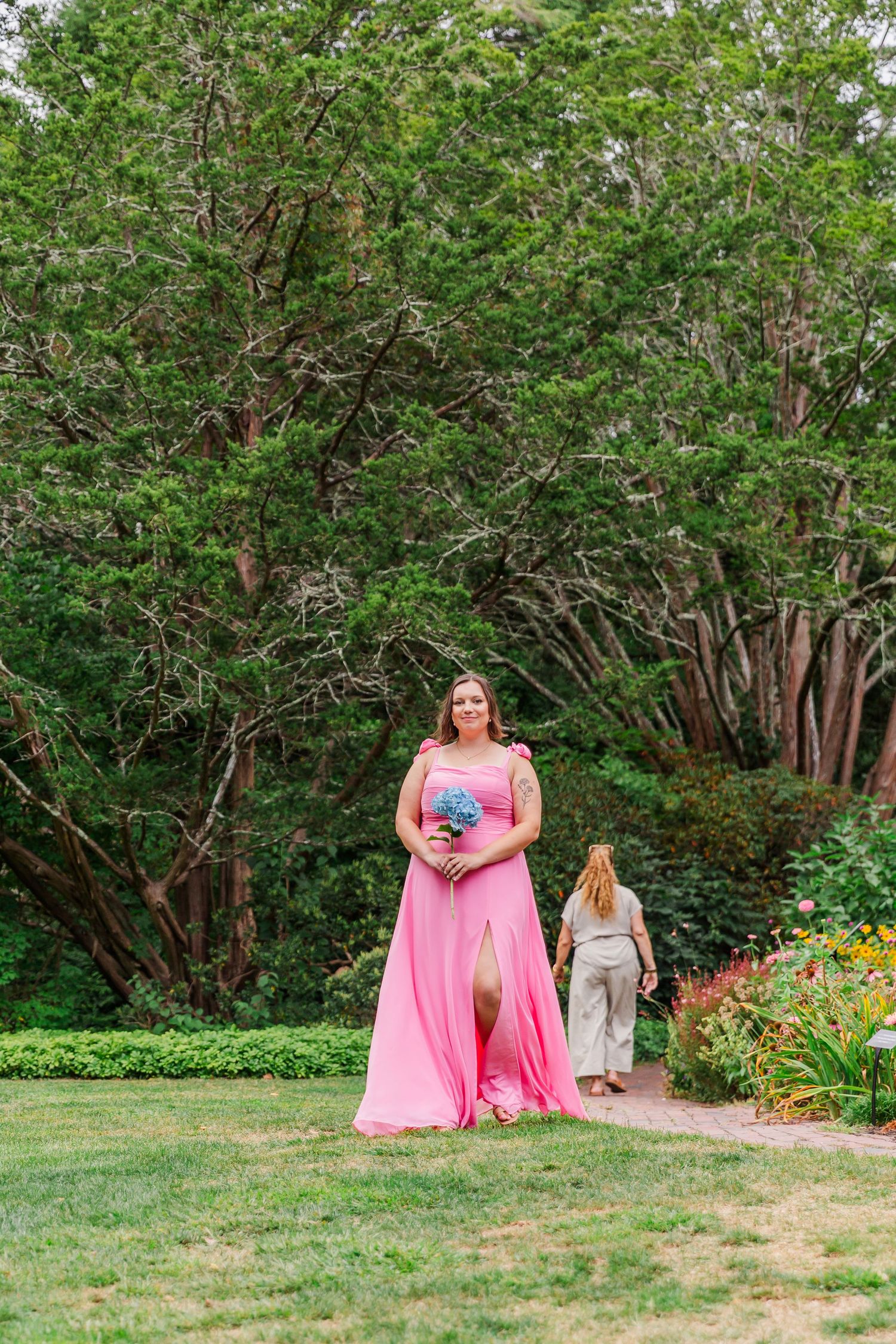 A bridesmaid in a flowing pink gown holds a bouquet while walking through a lush garden path.