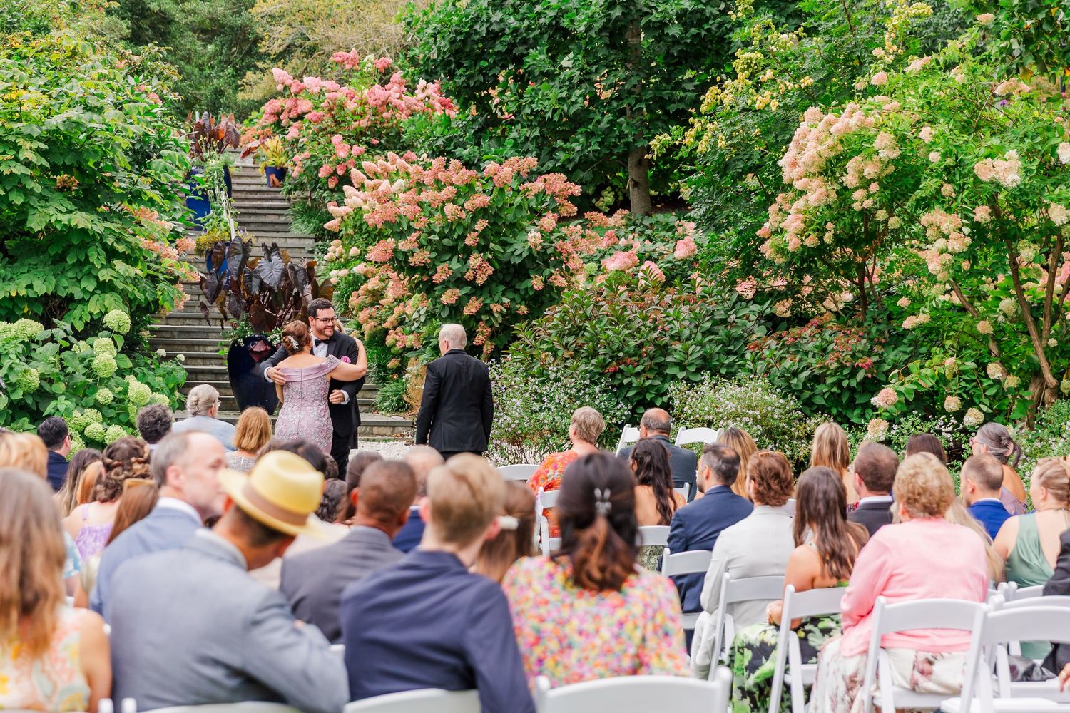 Guests seated at an outdoor garden wedding ceremony surrounded by blooming pink roses and lush greenery.
