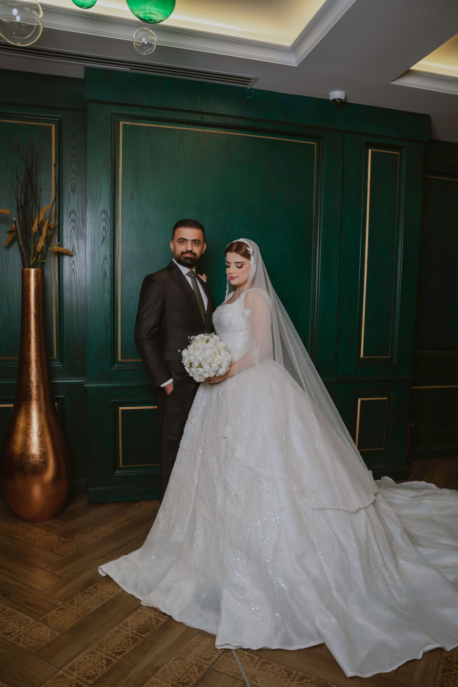 A bride in a flowing white wedding gown with a long veil poses elegantly in an emerald green paneled room.