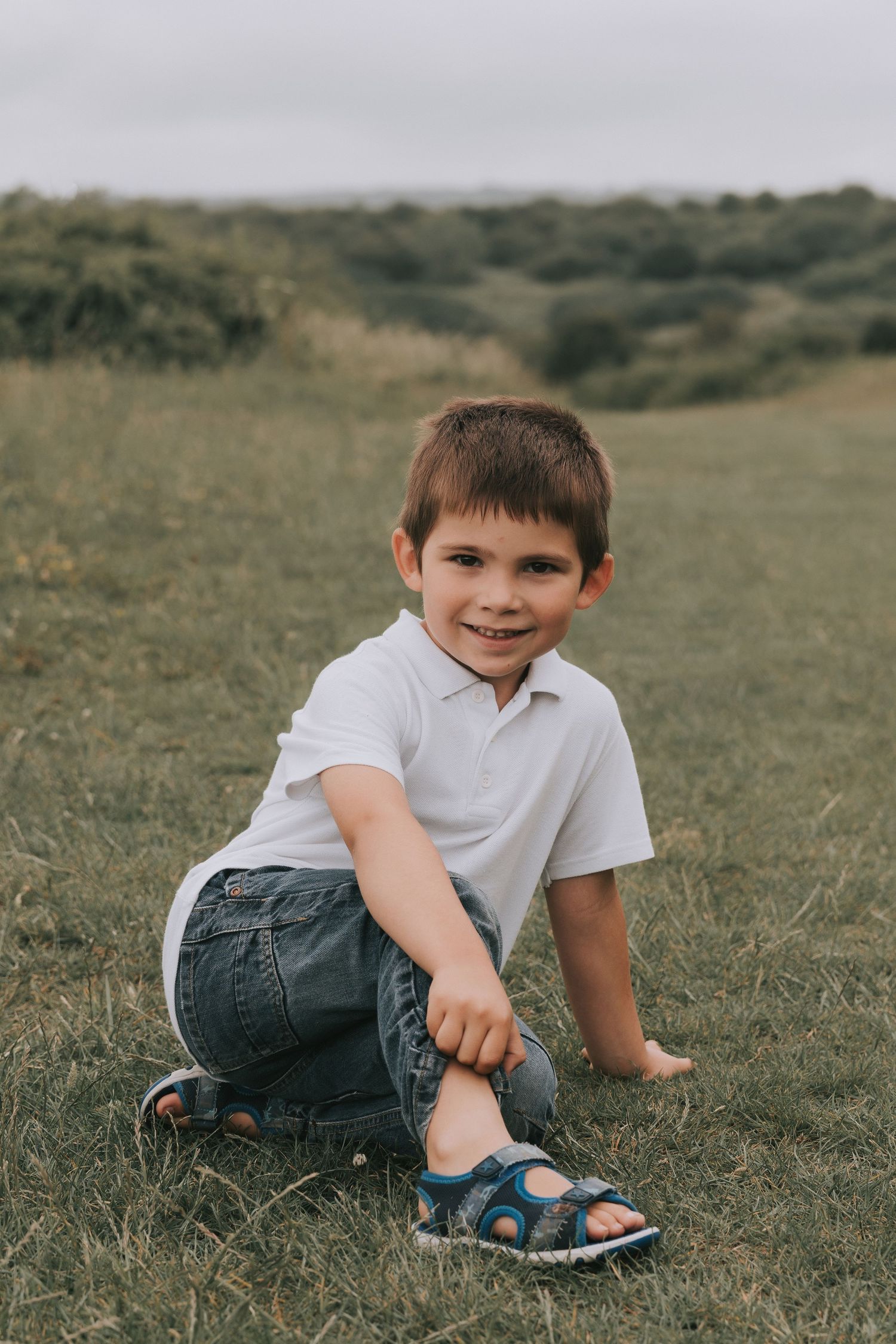 Young child in white polo shirt and jeans sitting on grassy hillside.