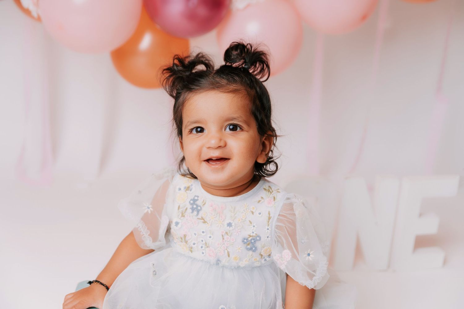 Baby in white dress sitting among pink and orange balloons at birthday photoshoot.