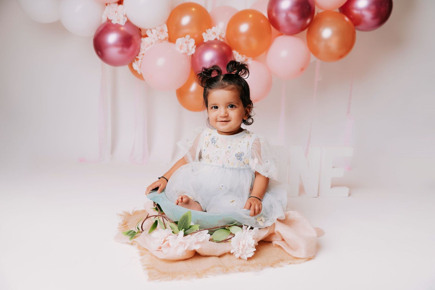 Baby celebrating first birthday with cake and balloon decorations in pink and orange.