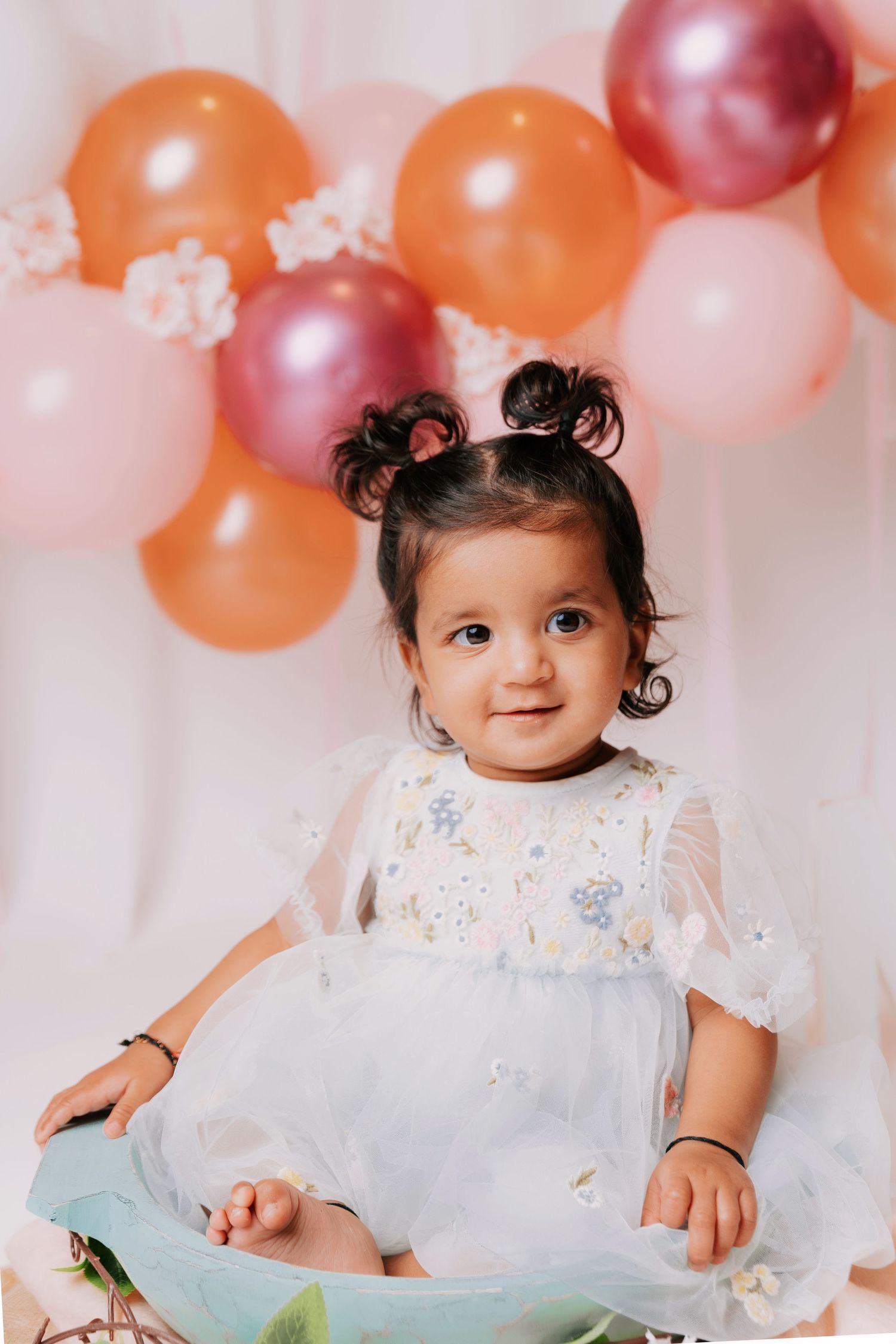 Baby in white dress sitting with birthday cake surrounded by pink and orange balloons.