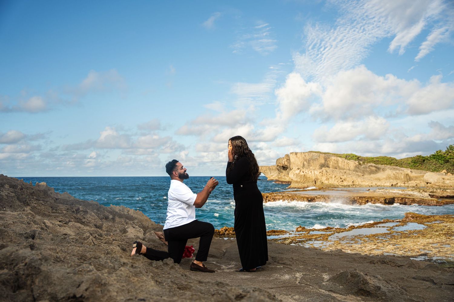 Two people sit together on rocky coastal cliffs overlooking the ocean waves and rugged shoreline at sunset.