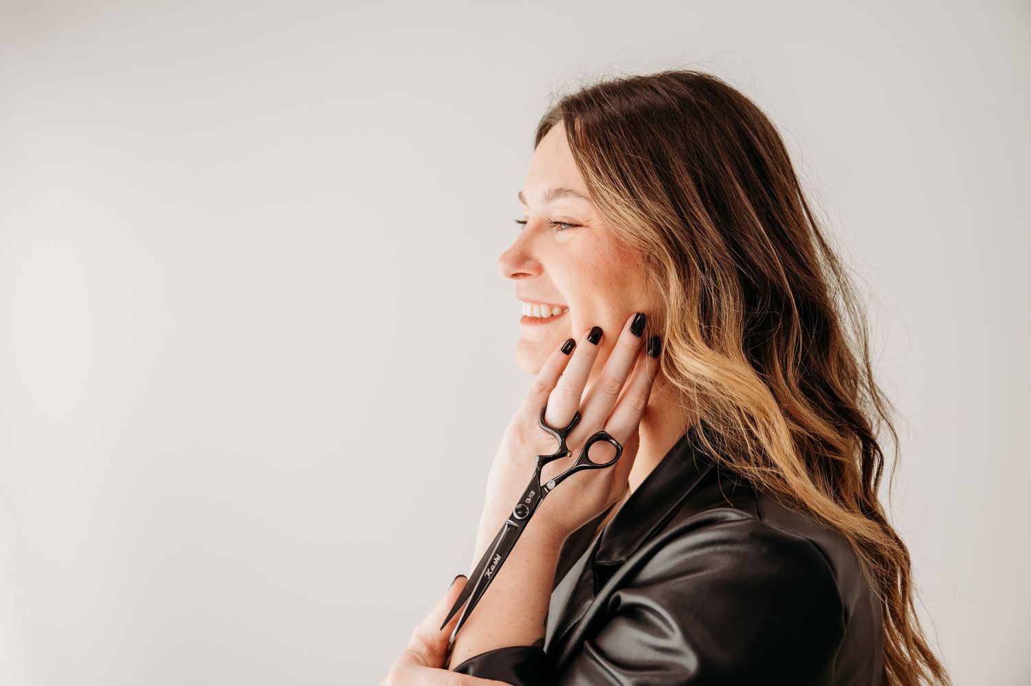 A smiling brunette with highlights gently touches her face while holding her haircutting shears for branding photos.
