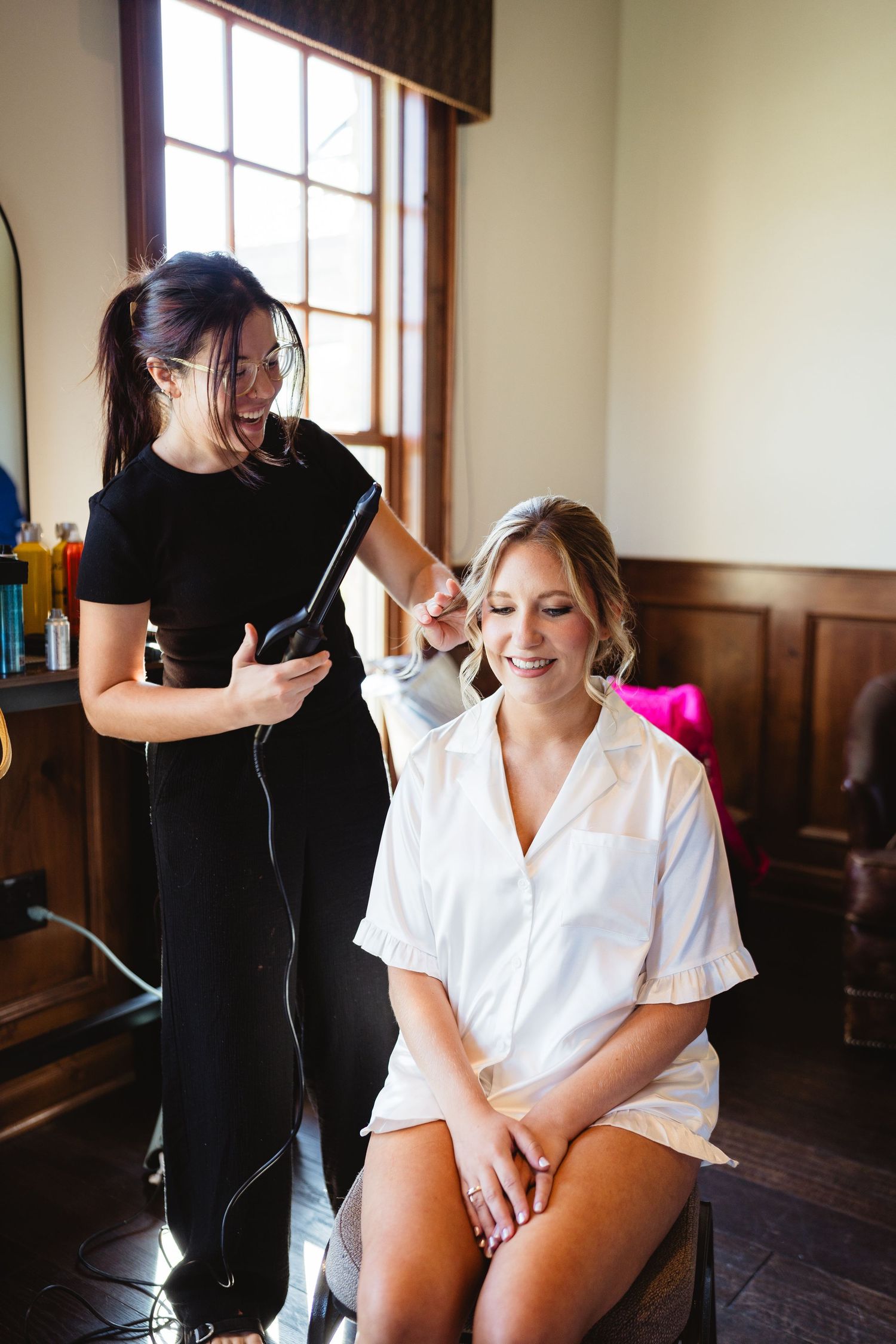 Hairstylist working on client's hair in a room with wooden paneled walls and large windows.