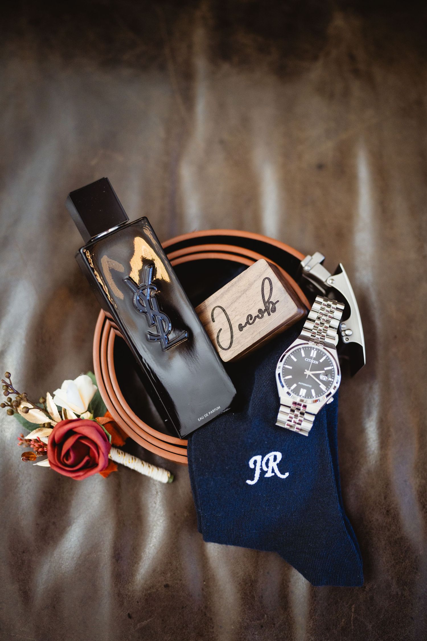Wedding accessories including a watch, leather goods, and navy blue tie arranged artistically on a textured background.