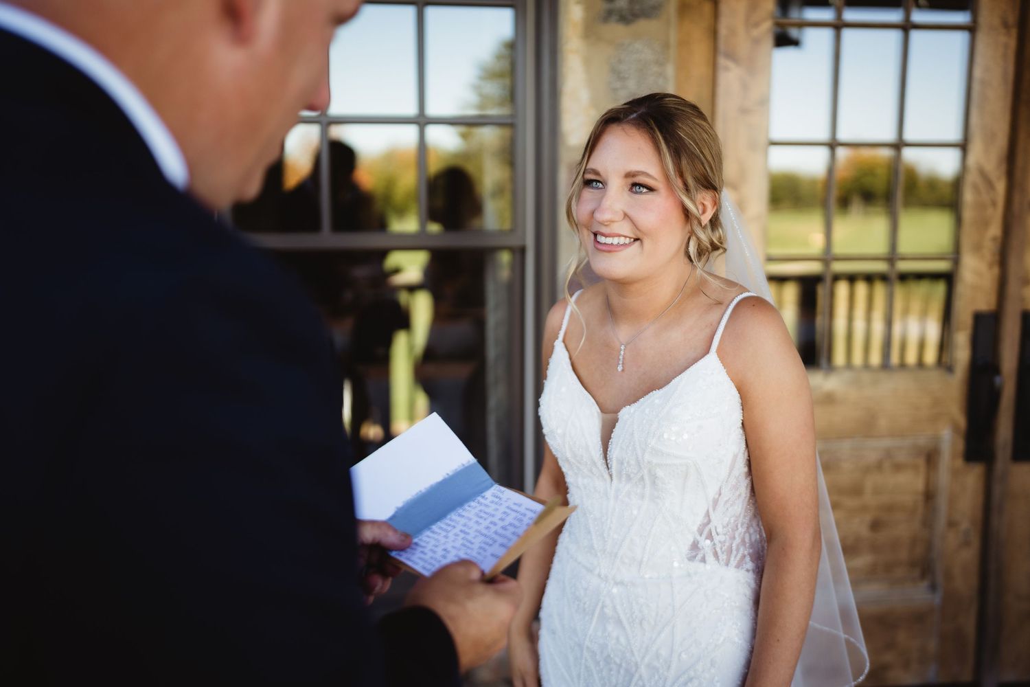 A bride in a white lace wedding dress stands smiling near a window during a wedding ceremony.