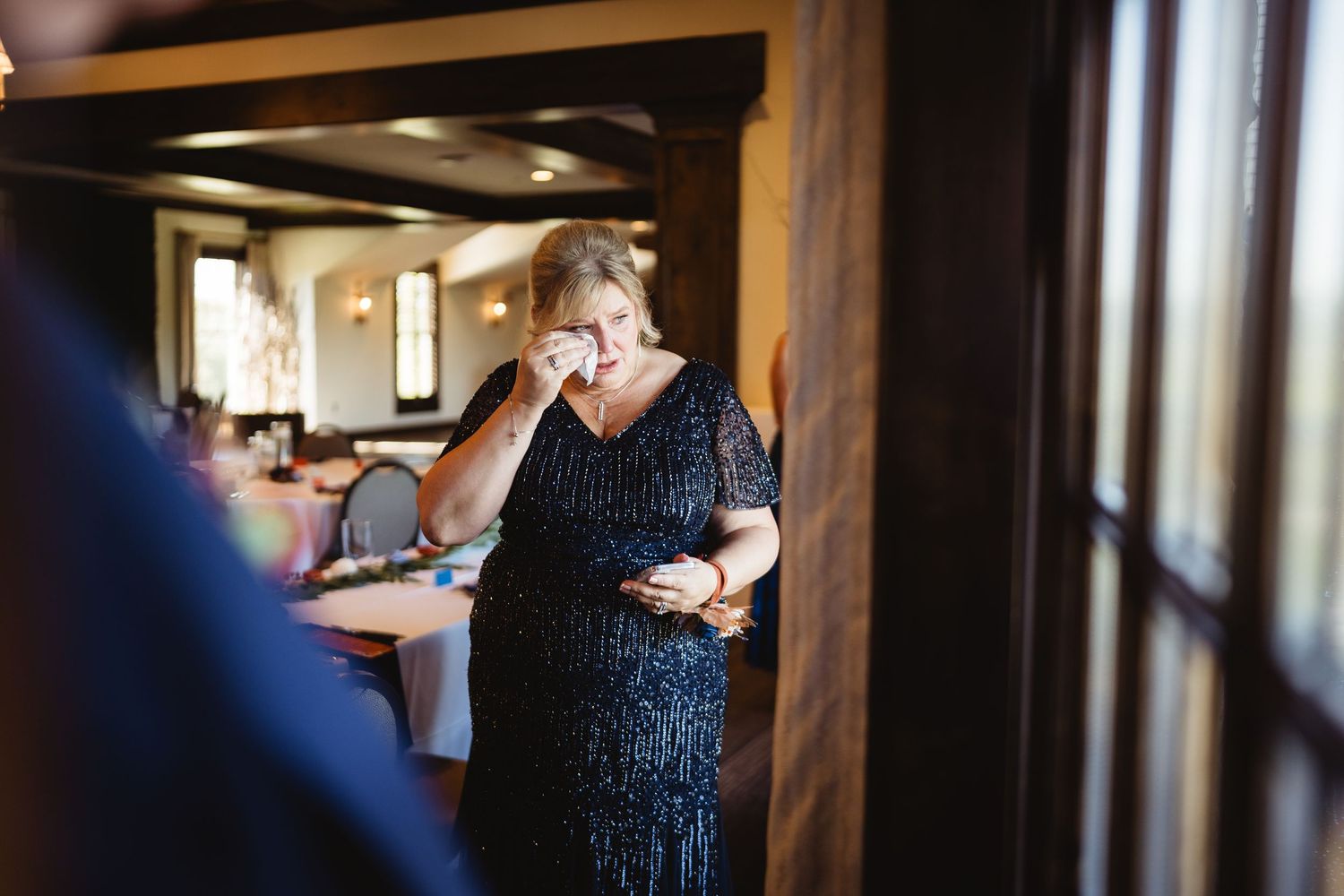 A person in a dark sequined dress stands in a sunlit hallway near wooden beams and white walls.