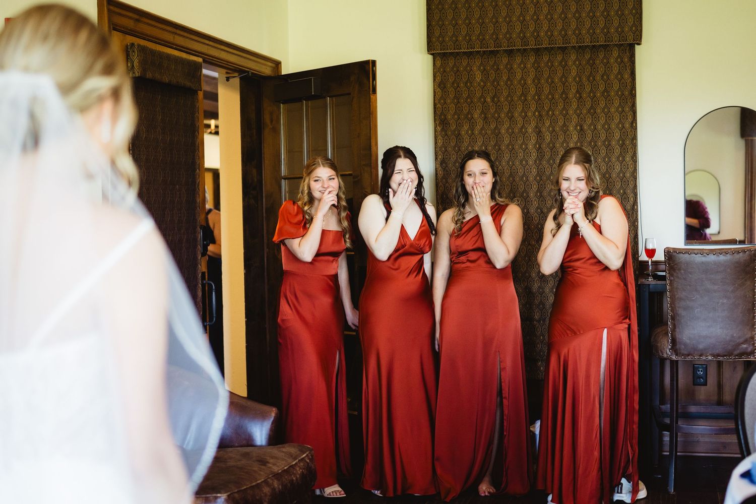A group of bridesmaids in red dresses react with joy and surprise during a wedding day moment.