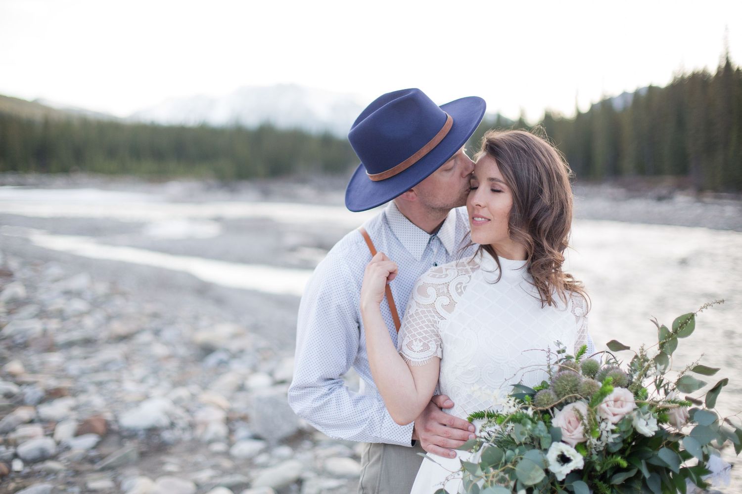 Romantic wedding couple sharing an intimate moment by the river while holding a floral bouquet in the mountains.