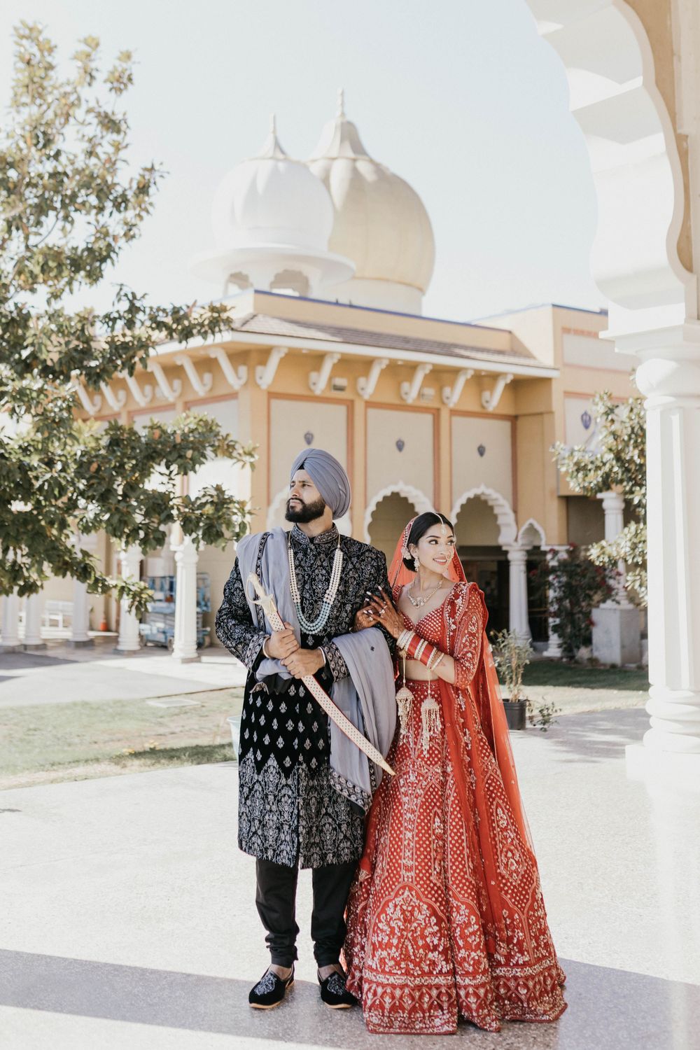 A couple poses in traditional wedding attire outside a white temple with Indian architectural features.