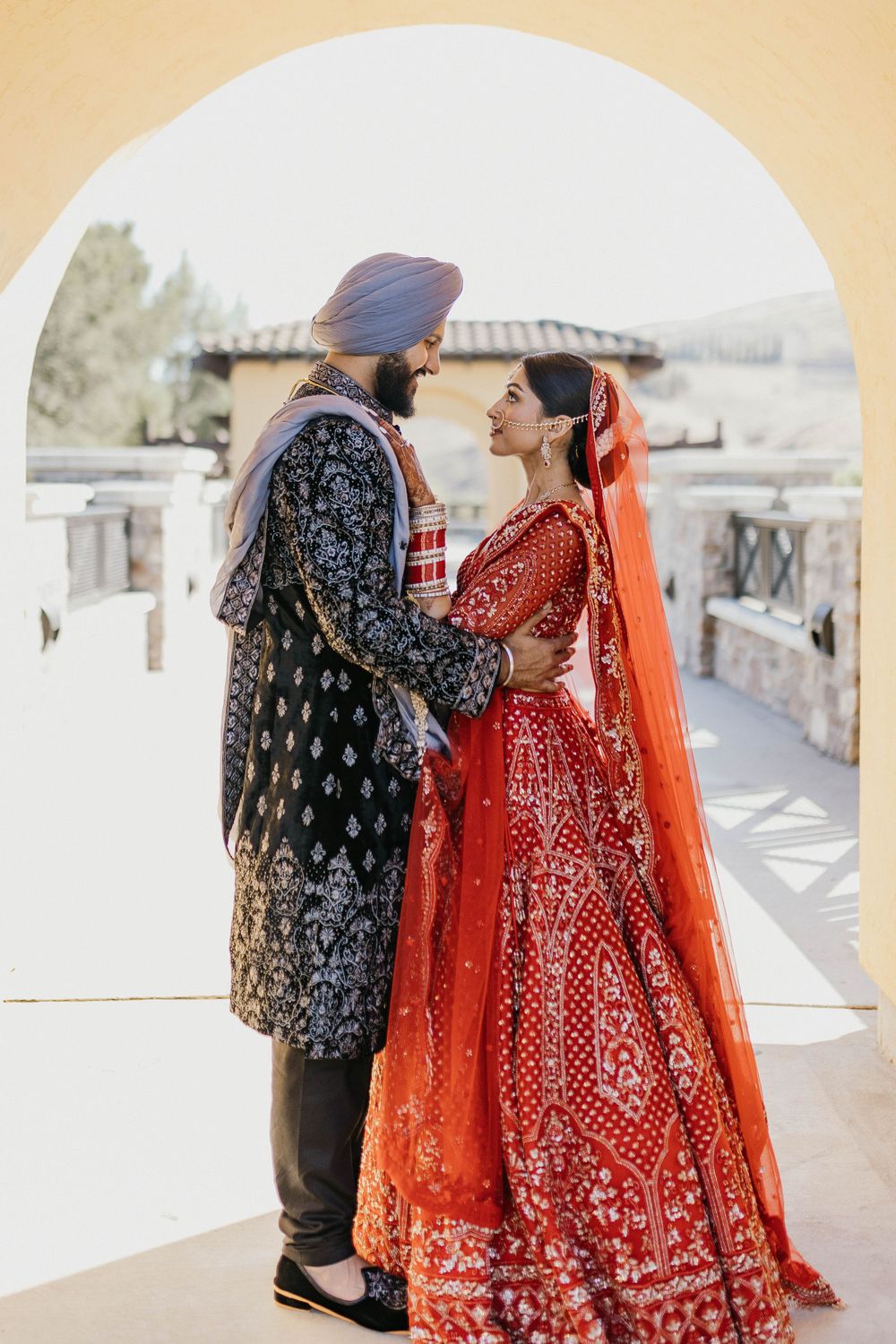 A couple in traditional Indian wedding attire with a vibrant red lehenga and black patterned sherwani walk outdoors.