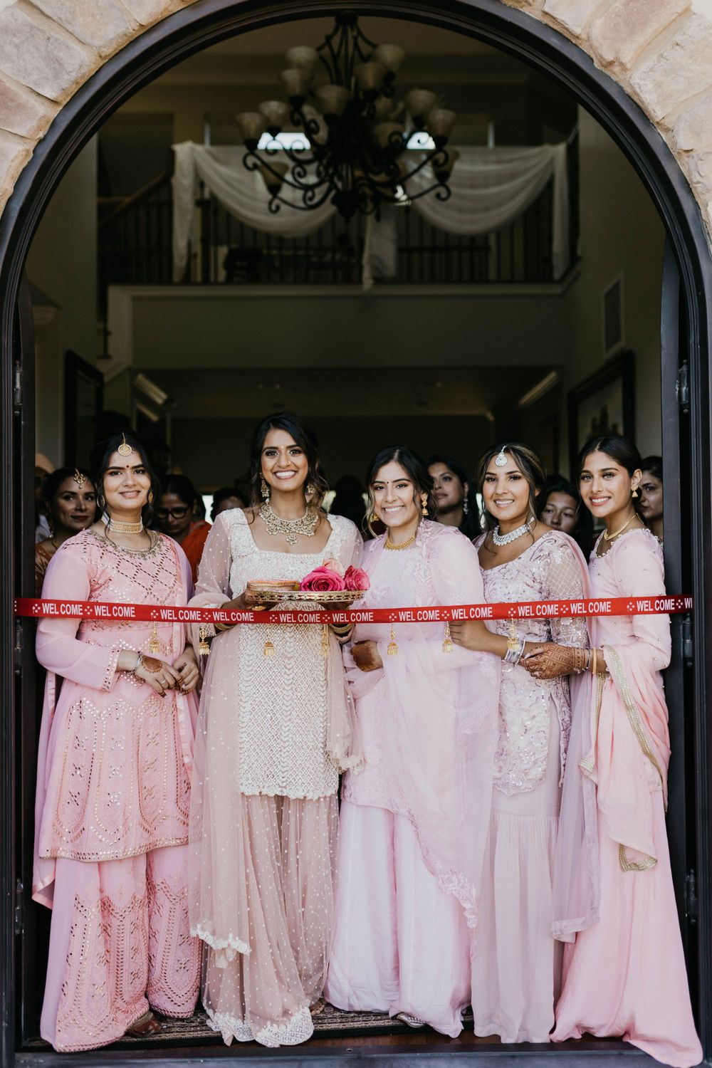 Wedding party wearing coordinated pink and cream outfits poses under an arched doorway.
