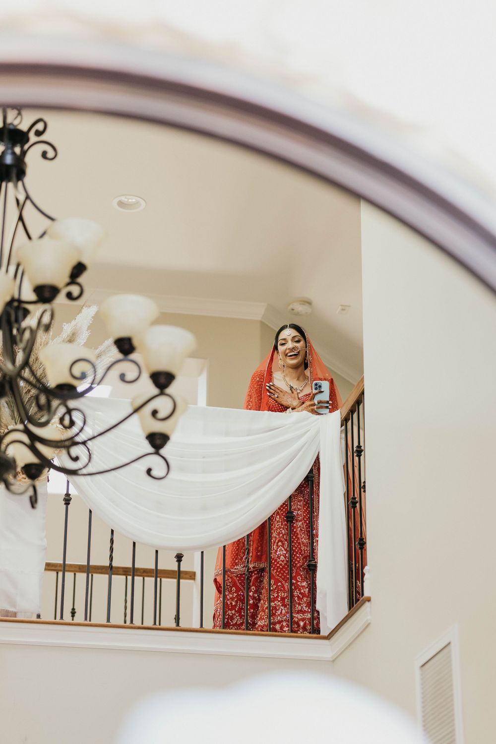 Bride in red lehenga poses on an elegant indoor balcony with white draping and chandelier.