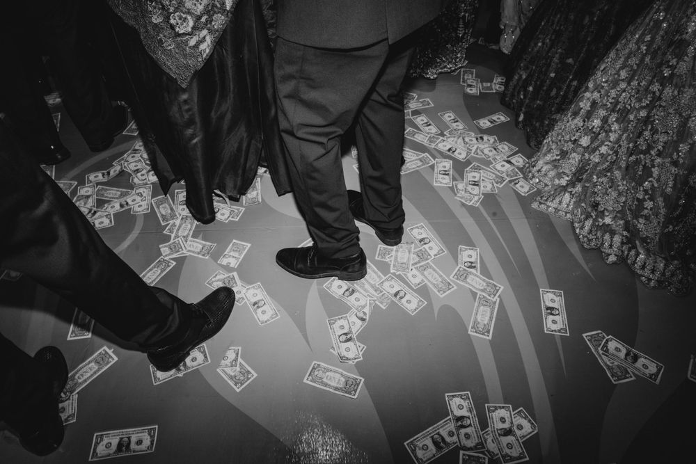 Black and white image of dollar bills scattered on a reflective floor with people's feet visible.
