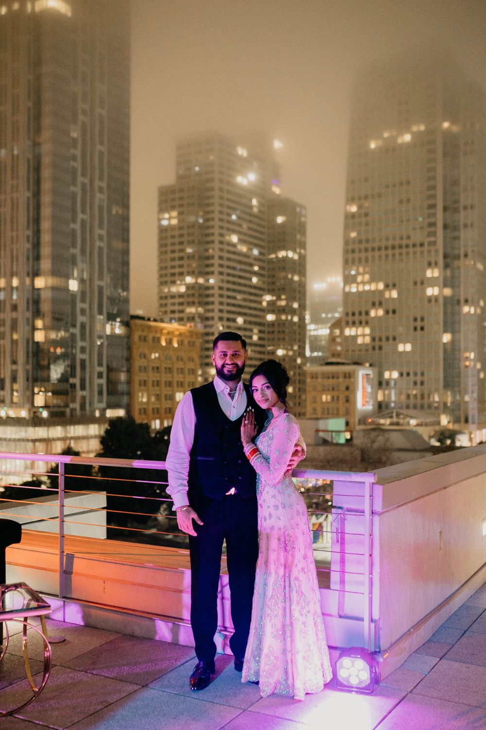 Couple in formal attire on a rooftop with foggy city skyline at night.