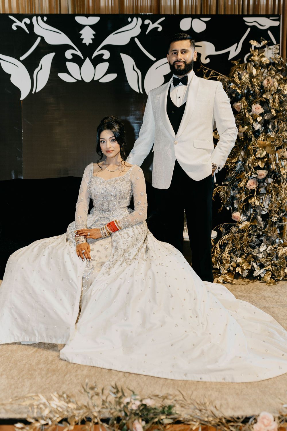 Wedding couple in white formal attire posing against black and white floral backdrop.