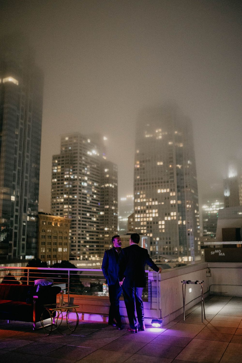 Foggy city skyline at night with purple lights illuminating figures on a rooftop.