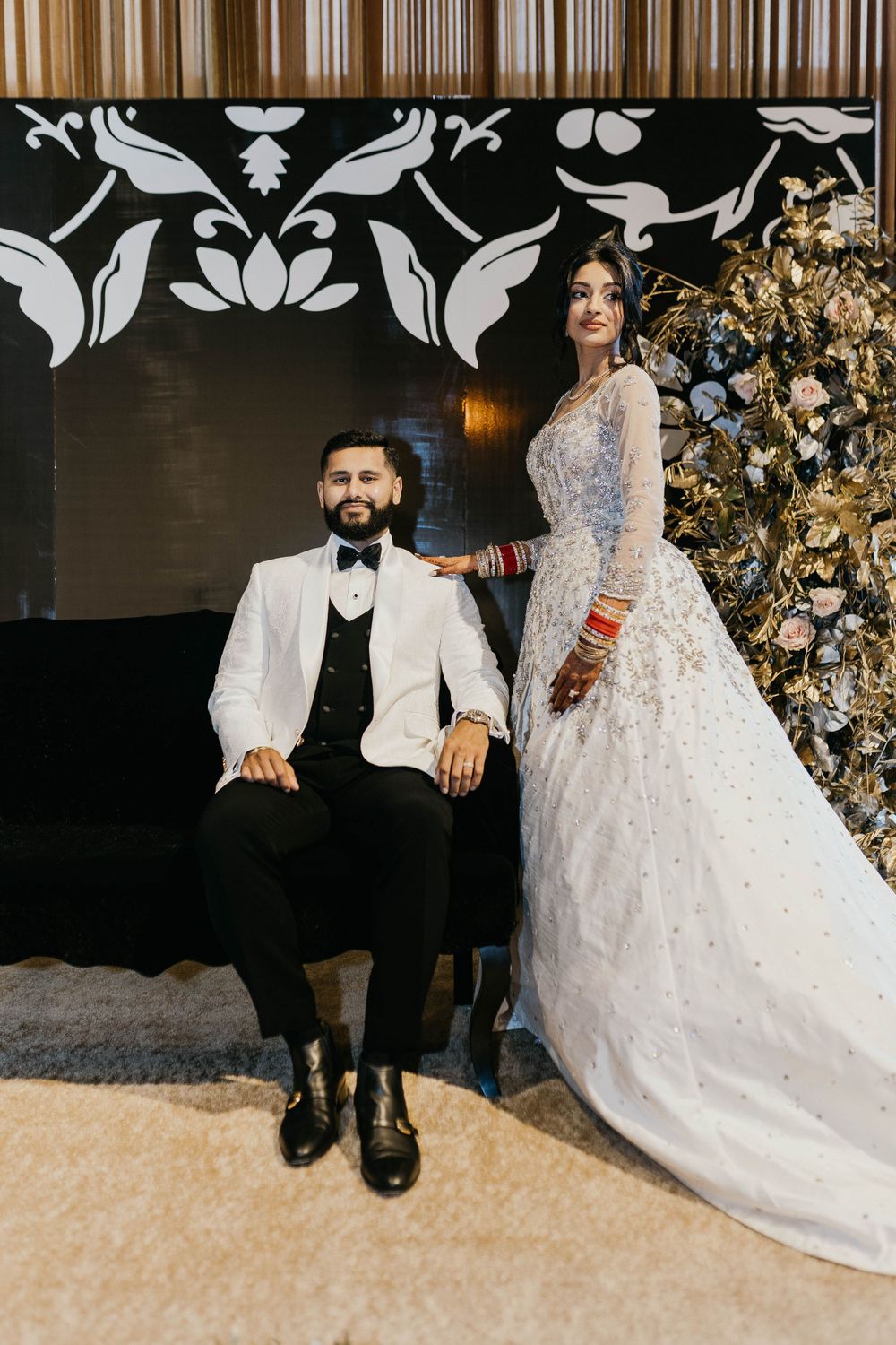 Couple in elegant wedding attire seated against decorative black and white backdrop.