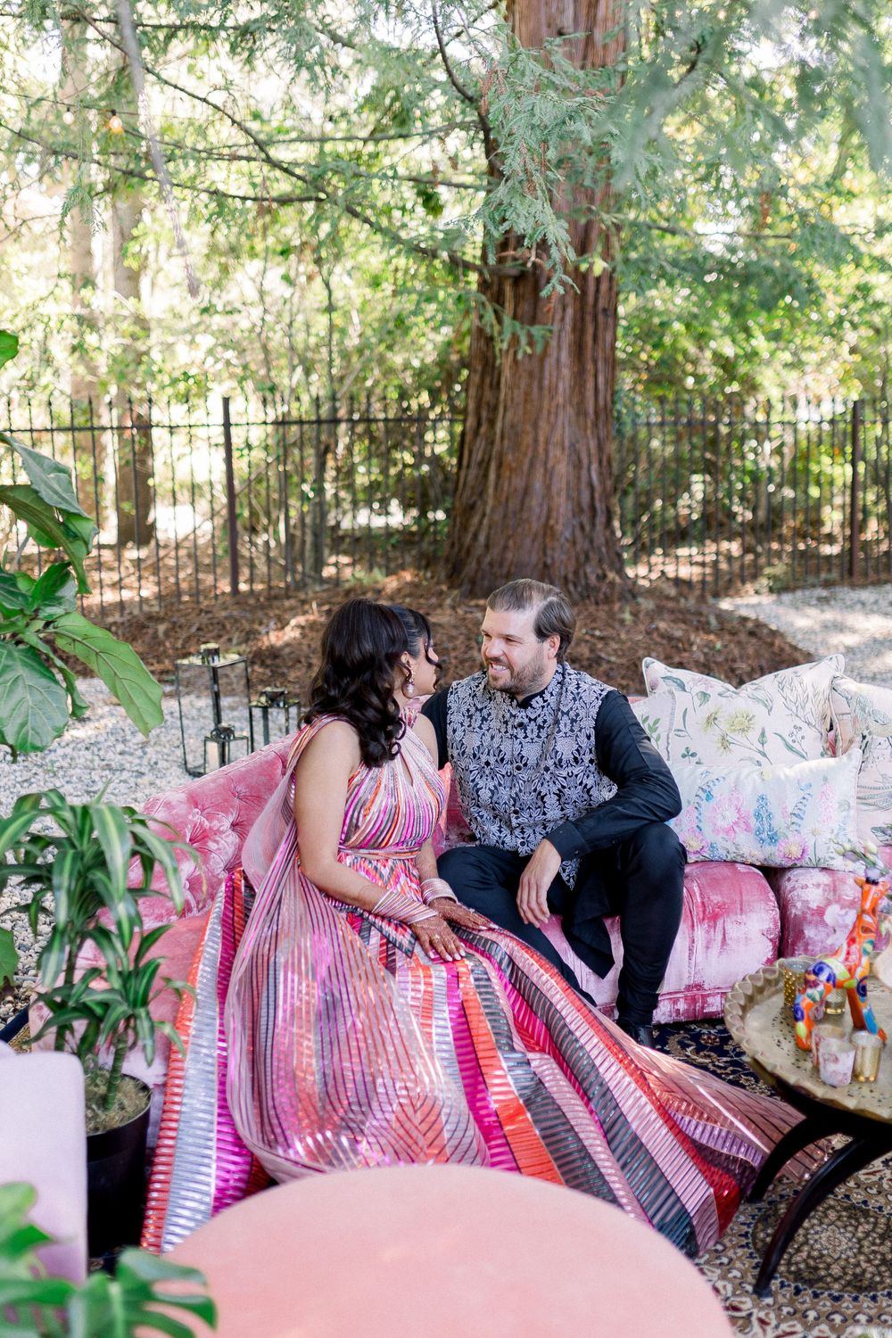 Romantic outdoor photoshoot with couple seated on pink pillows surrounded by trees and whimsical decor.