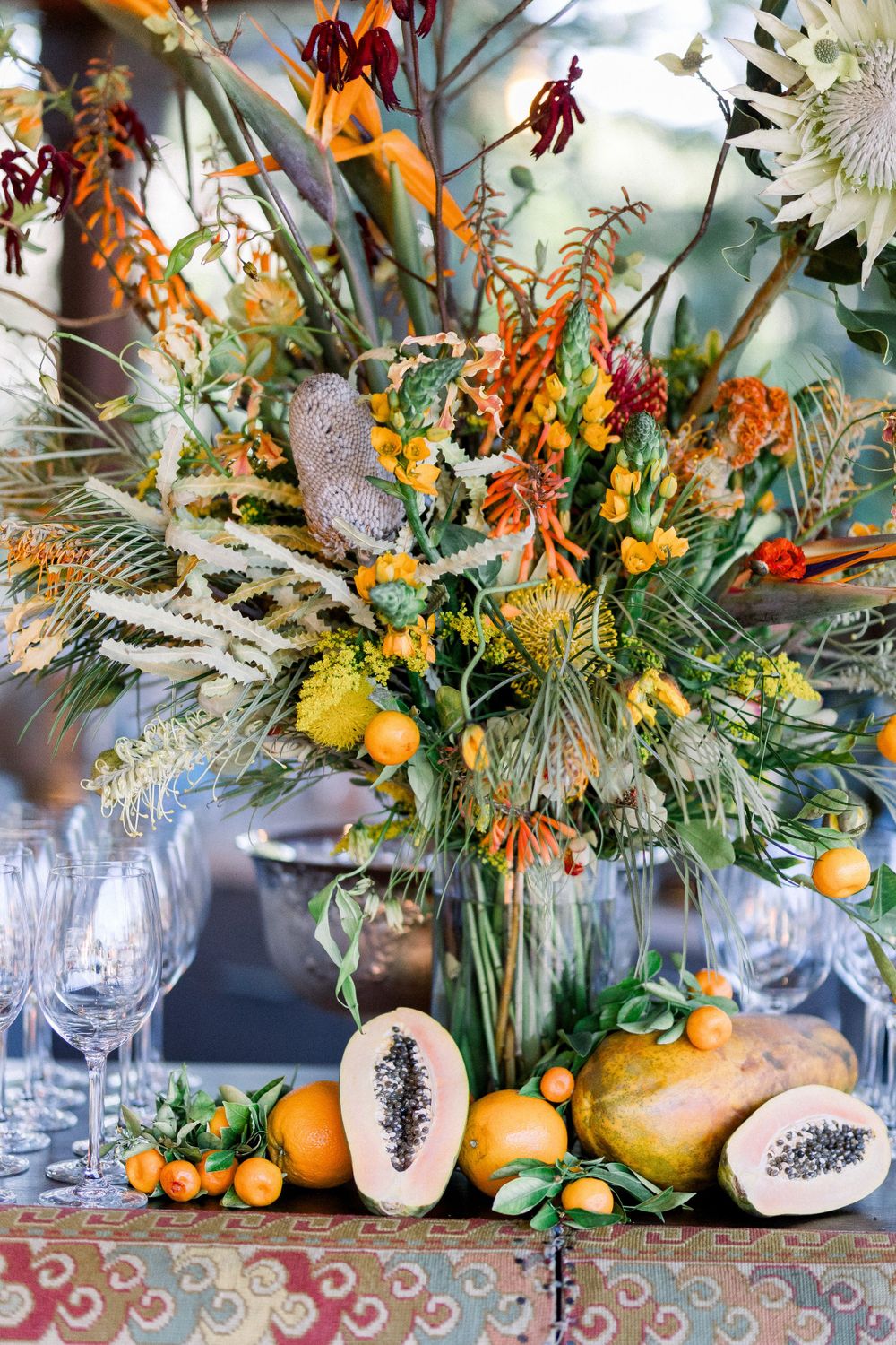 Vibrant fall centerpiece with orange flowers, dried elements, and citrus fruits arranged with glassware.