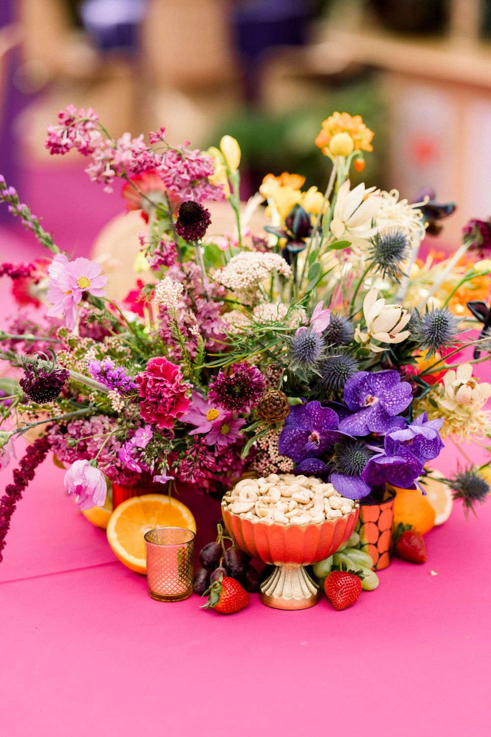 Vibrant floral centerpiece with purple and pink blooms alongside citrus and berries on pink surface.