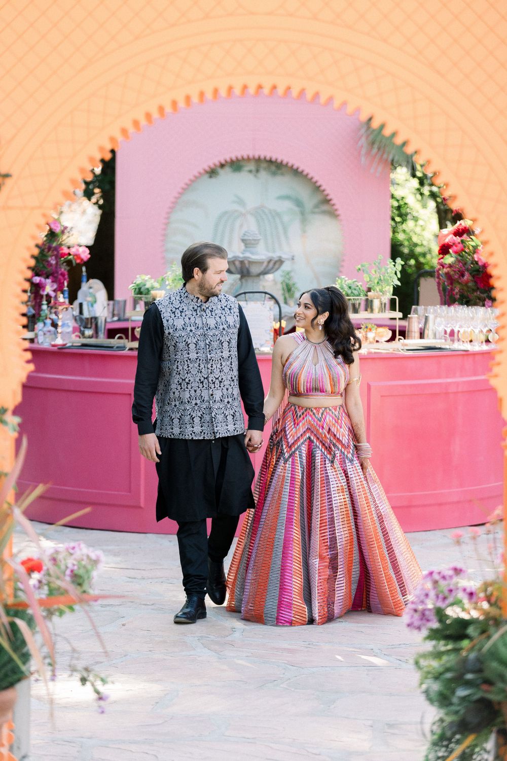 Couple in festive attire standing in front of a pink circular bar under decorative arch.