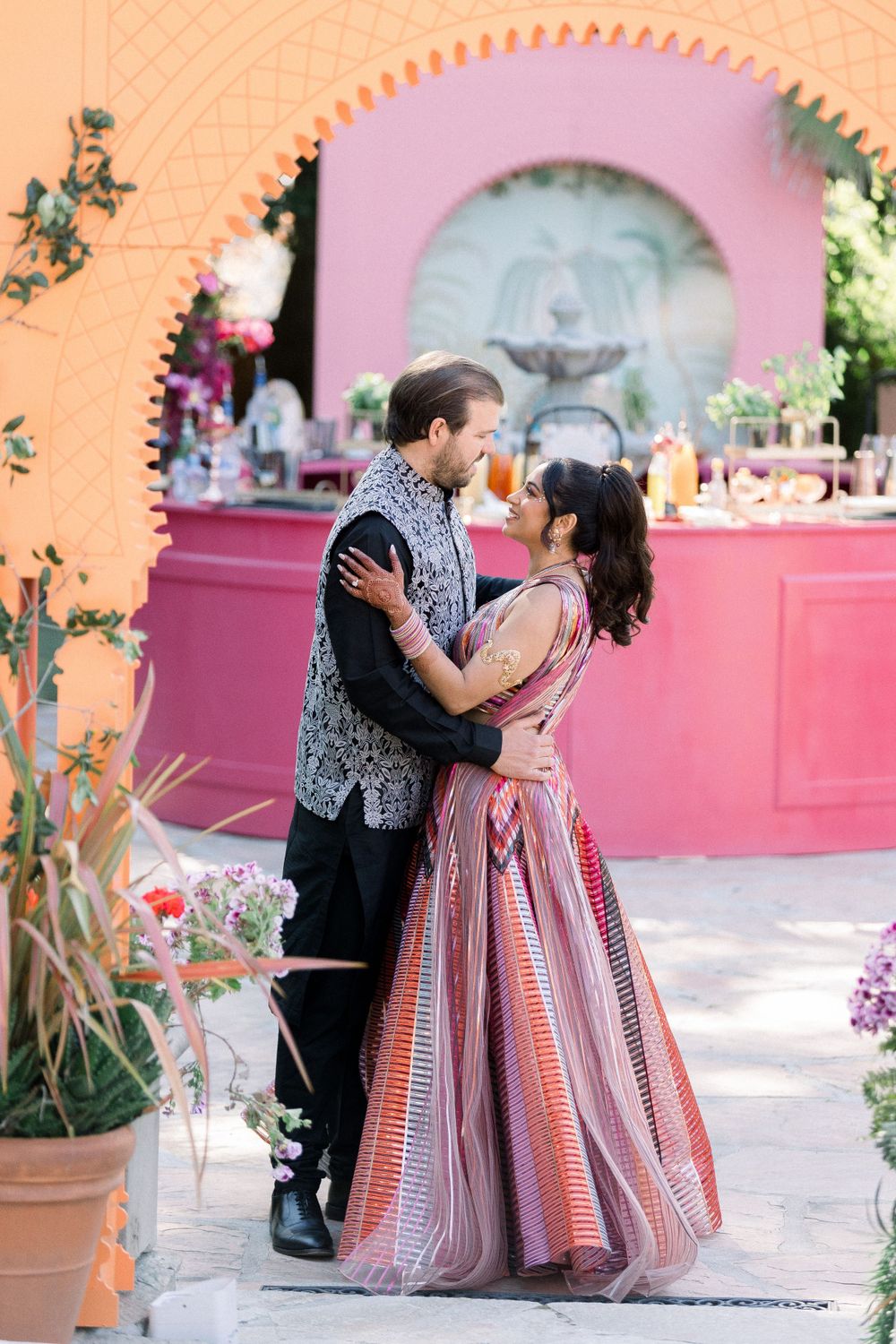 Couple embracing under peach archway with pink bar setting in background.