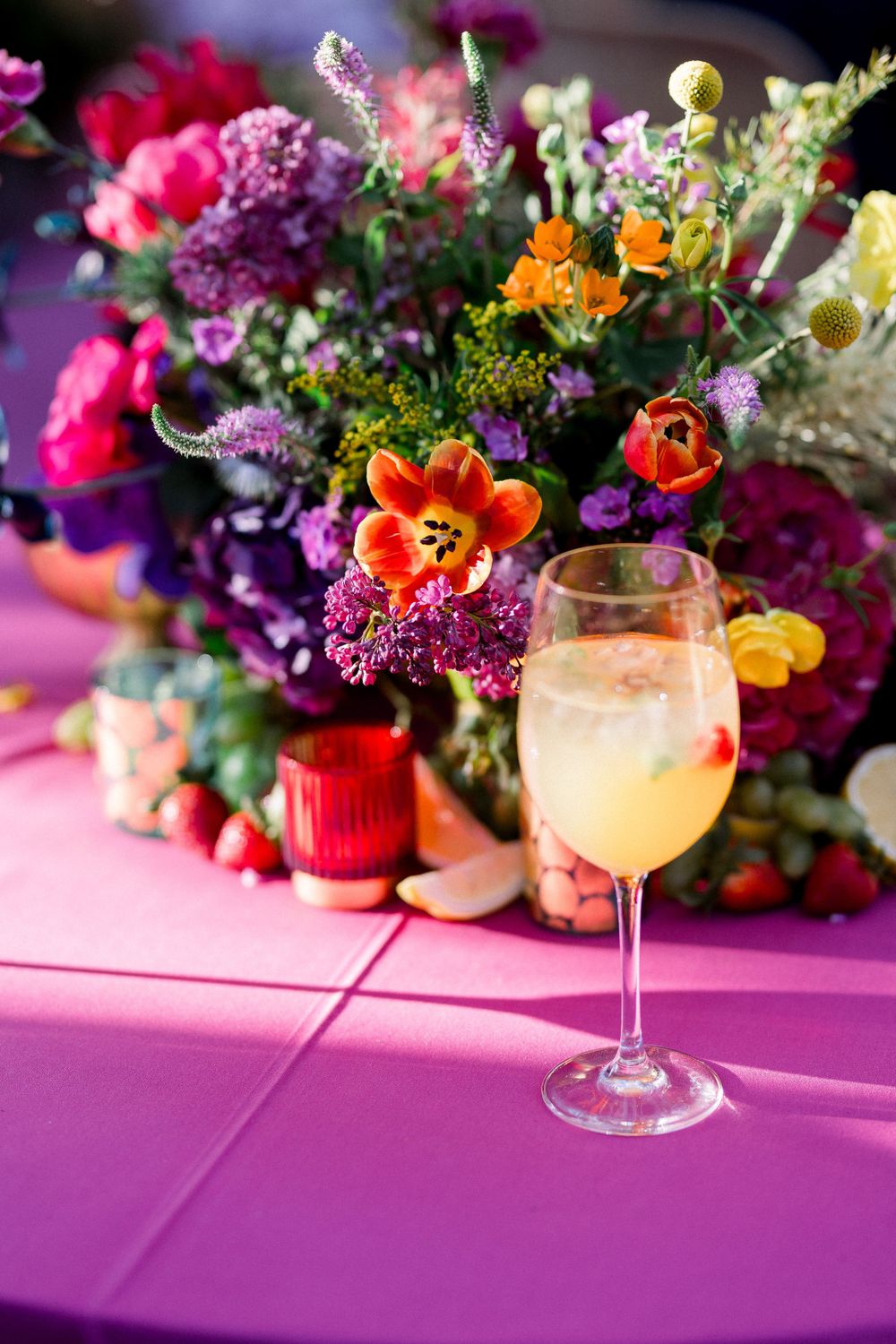Vibrant floral arrangement with orange and purple blooms next to a champagne glass on pink tablecloth.