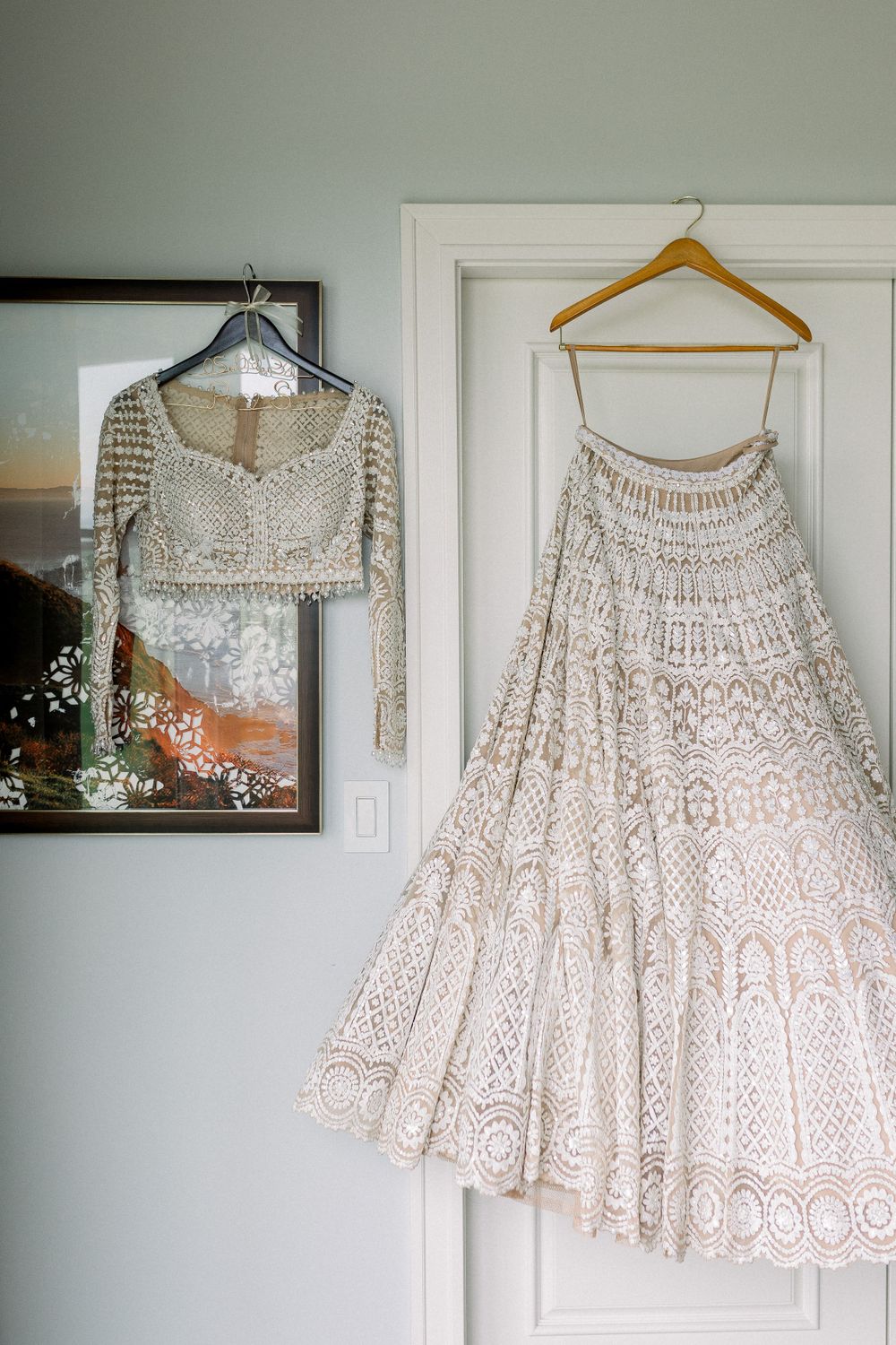 A collection of elegant white embroidered lehengas and wedding dresses displayed on hangers against a pale wall.