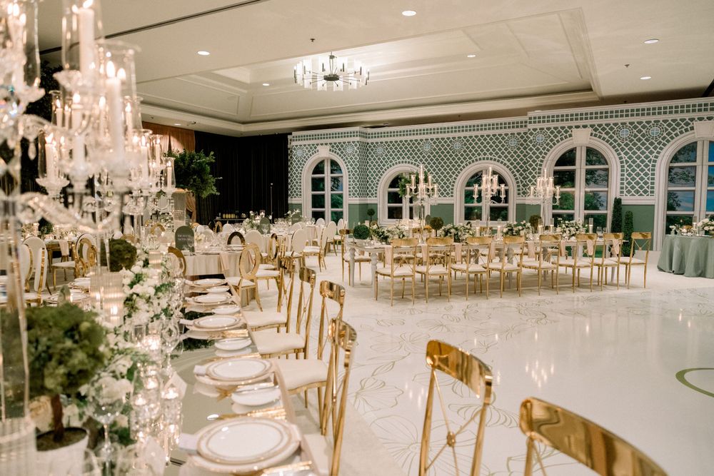 Luxurious ballroom setup with crystal chandeliers, arched windows, and gold chairs around white-clothed tables.