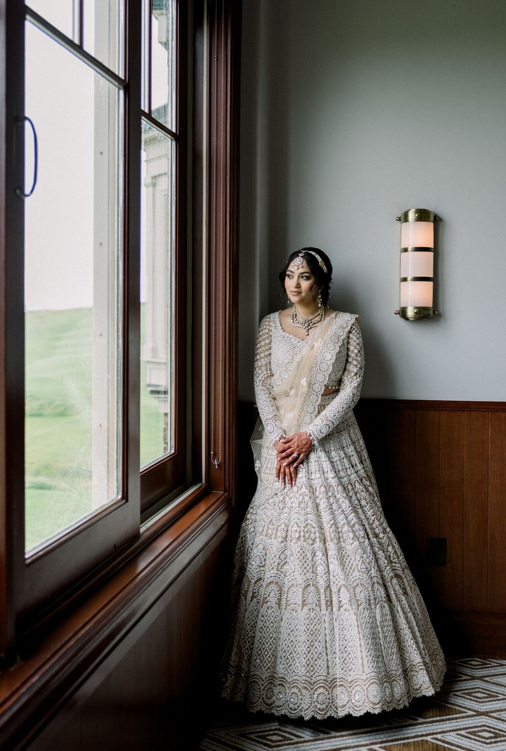Bride in an ornate lace gown poses by large windows in natural light.
