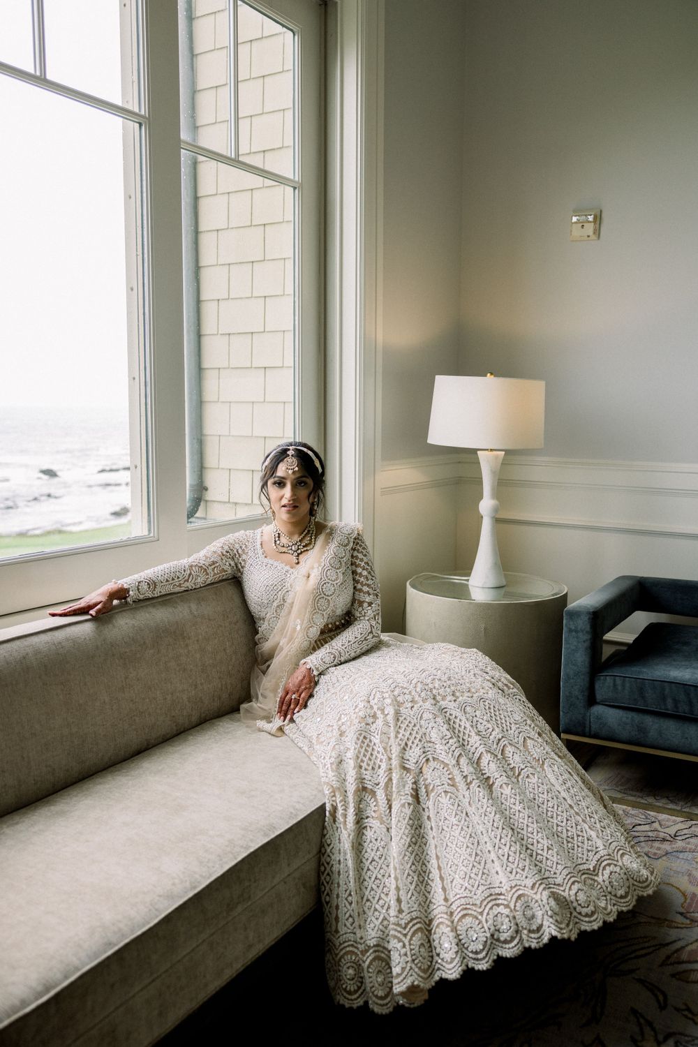 Bridal portrait in a sunlit room with lace dress arranged on a gray couch by window.
