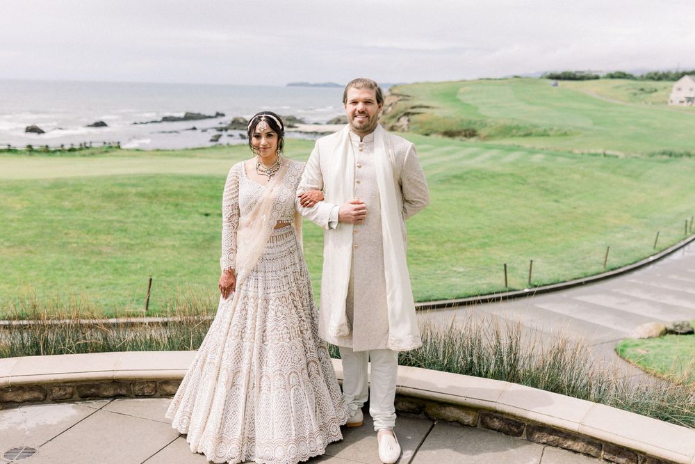 Elegant couple in cream-colored wedding attire walking along a coastal pathway.